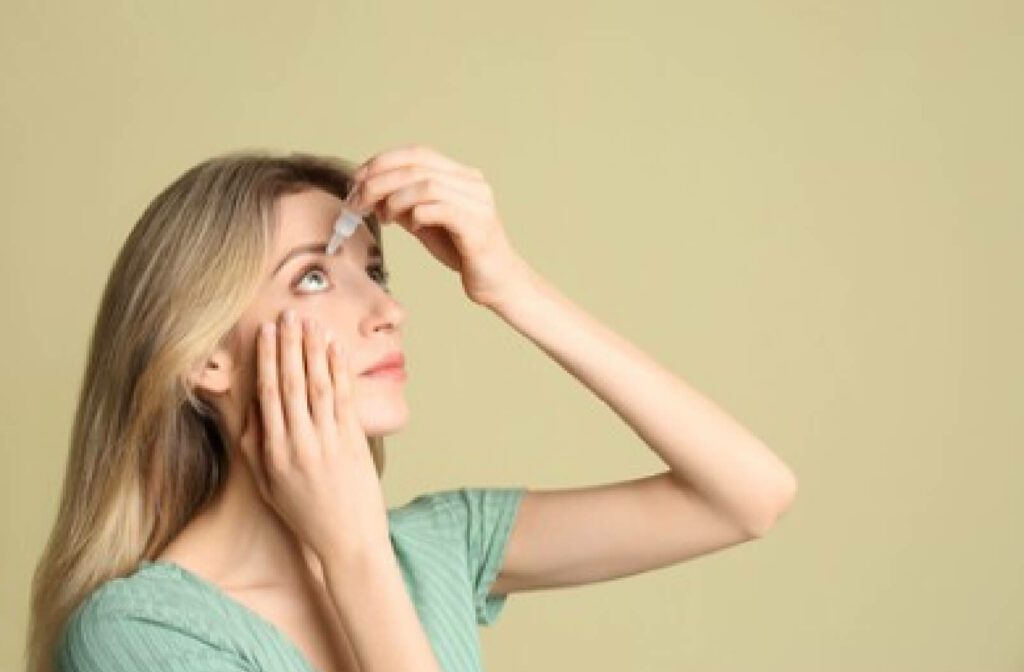 A young woman is holding a bottle of eye drops close to her right eye and pours some drops to lessen the discomfort because of dry eyes.