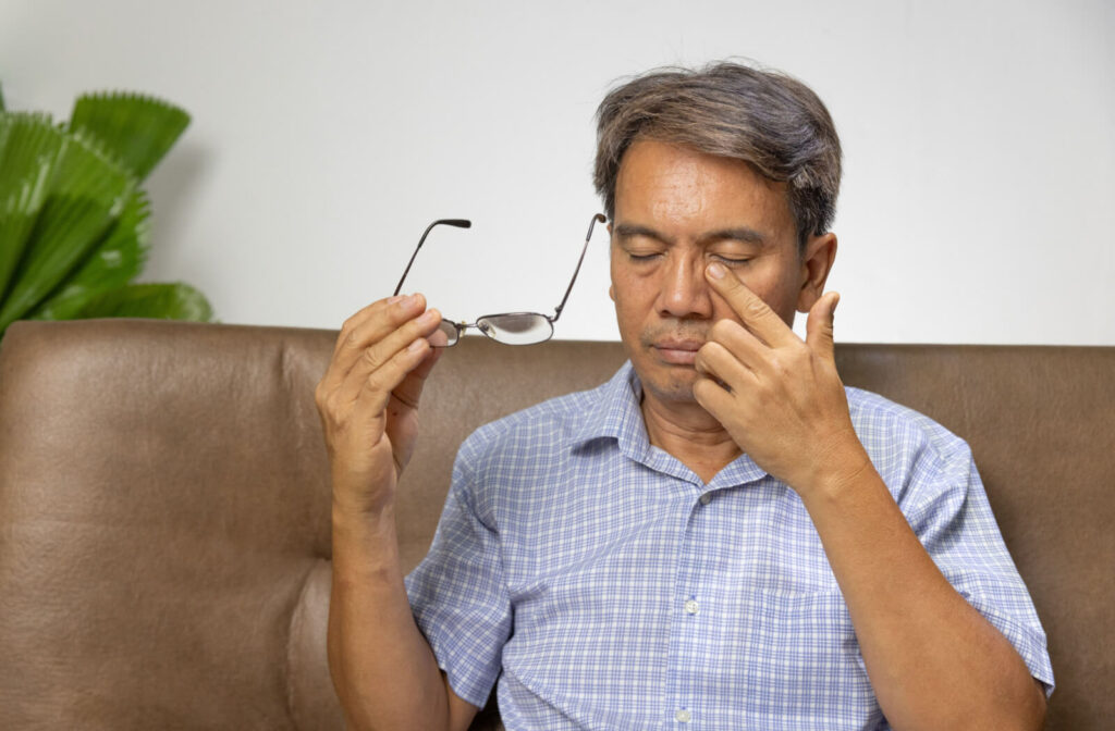 A senior man sitting on a couch and holding his glasses in his right hand as he rubs his left eyes with his left hand.