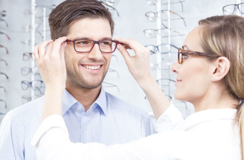 A young man trying on glasses at an optical store while being assisted by an optician or optometrist.