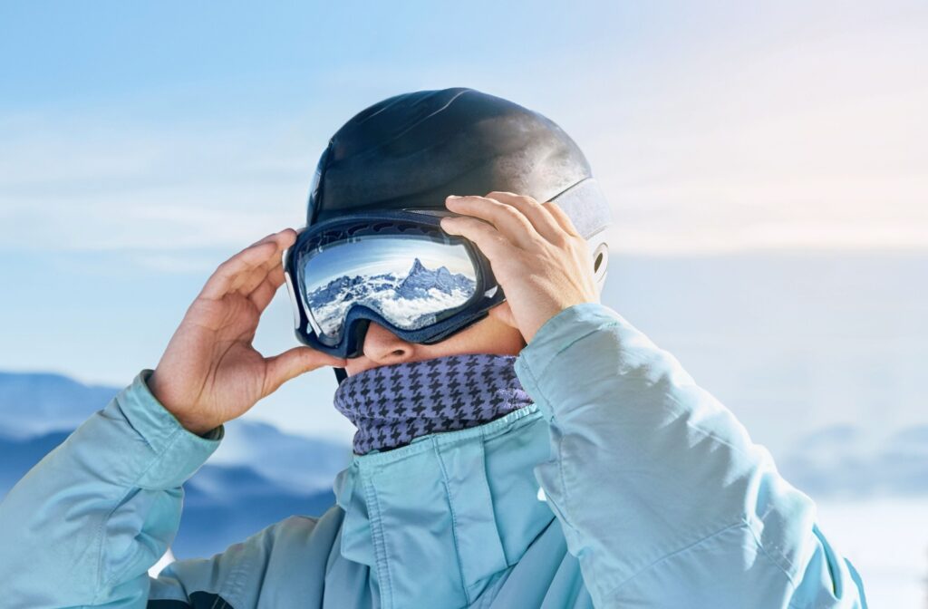 A skier secures protective goggles over their eyes to protect them from cold winds. A mountain range reflects in the goggles