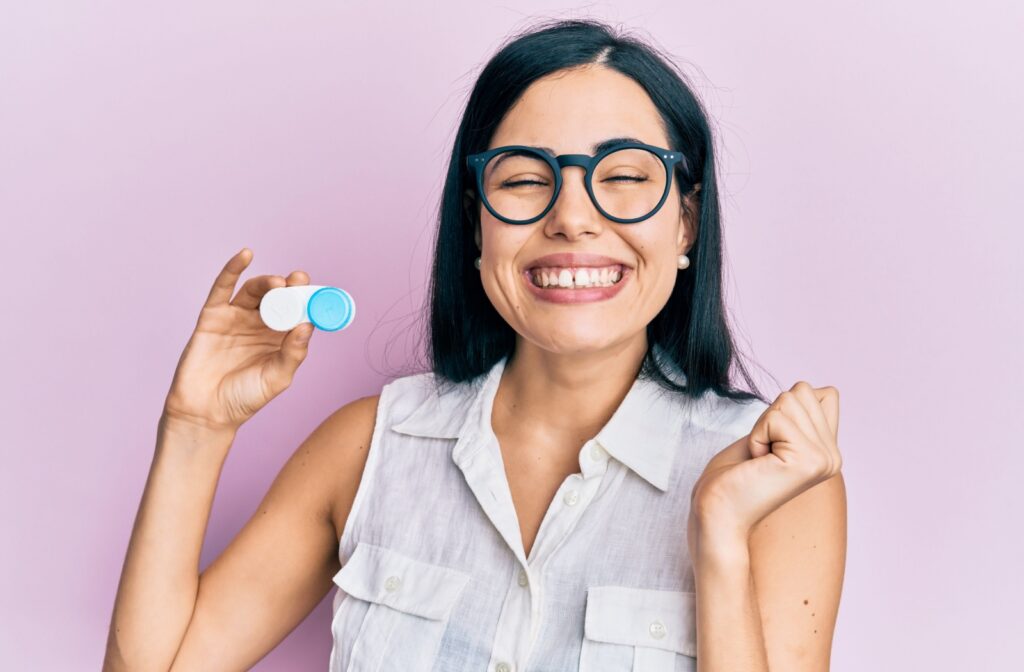 Against a pink backdrop, a grinning adult holds up contact lenses, excited to replace their eyeglasses in day-to-day use