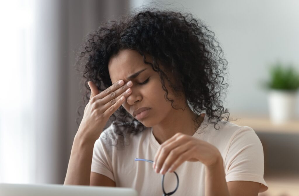 Young woman with curly black hair rubbing the bridge of their nose while holding eyeglasses in front of a computer screen.