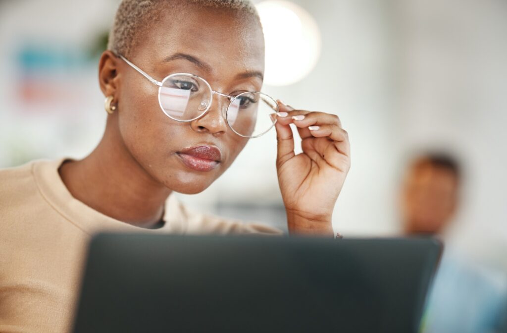 Woman wearing glasses looking at laptop screen while holding on to frame of glasses with her hand.