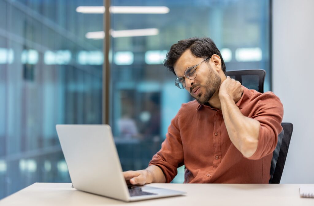 A person sitting at their desk on their laptop, rubbing their neck in pain in connection to digital eye strain